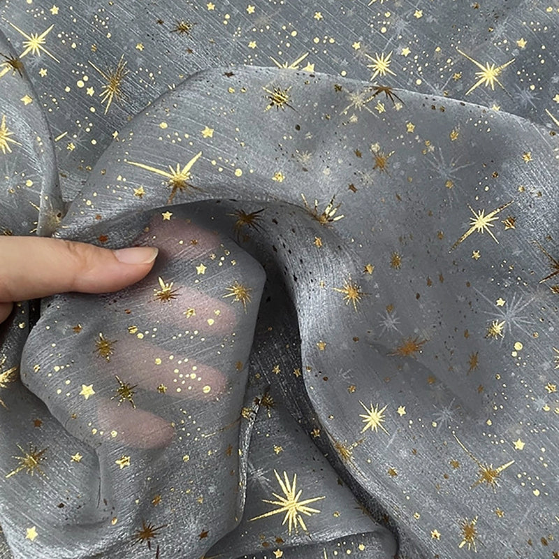 Une main tient le "Tissu organza étoilé au mètre" de Longan Craft France, un tissu transparent et argenté avec des motifs d'étoiles et de points dorés, parfait pour les robes de mariée et les costumes. L'organza semi-transparent laisse apparaître les doigts de la personne à travers lui.