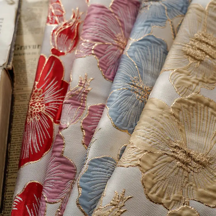 Plusieurs rouleaux de Longan Craft France Tissu brocart floral 3D en fil d'or, un élégant tissu jacquard d'ameublement aux vibrants motifs de brocart rouge, rose, bleu et beige, sont présentés côte à côte. Un journal et un livre apparaissent sur la gauche.