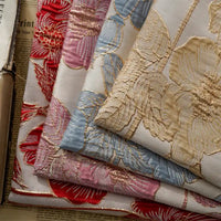 Quatre pièces de Tissu brocart floral 3D en fil d'or de Longan Craft France, en jacquard rouge, rose, bleu et beige, sont empilées luxueusement avec un soupçon de papier journal en dessous - parfait pour la haute couture ou un décor élégant.