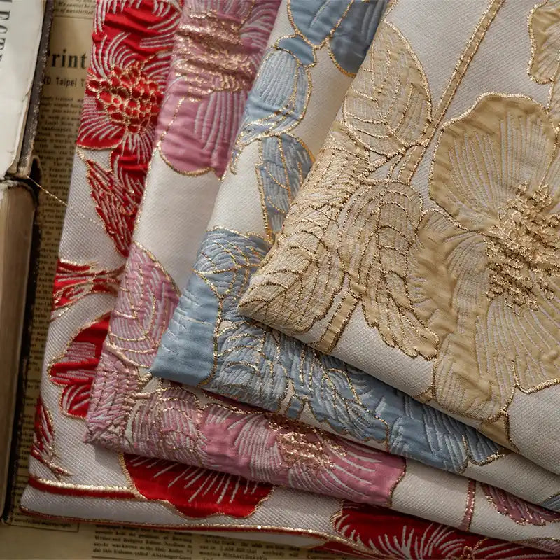 Quatre pièces de Tissu brocart floral 3D en fil d'or de Longan Craft France, en jacquard rouge, rose, bleu et beige, sont empilées luxueusement avec un soupçon de papier journal en dessous - parfait pour la haute couture ou un décor élégant.