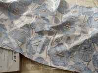 Un morceau de Tissu brocart floral 3D en fil d'or de Longan Craft France est posé sur des journaux ouverts, son élégant motif jacquard bleu, argenté et doré apportant une touche de luxe haute couture au-dessus des titres visibles en dessous.