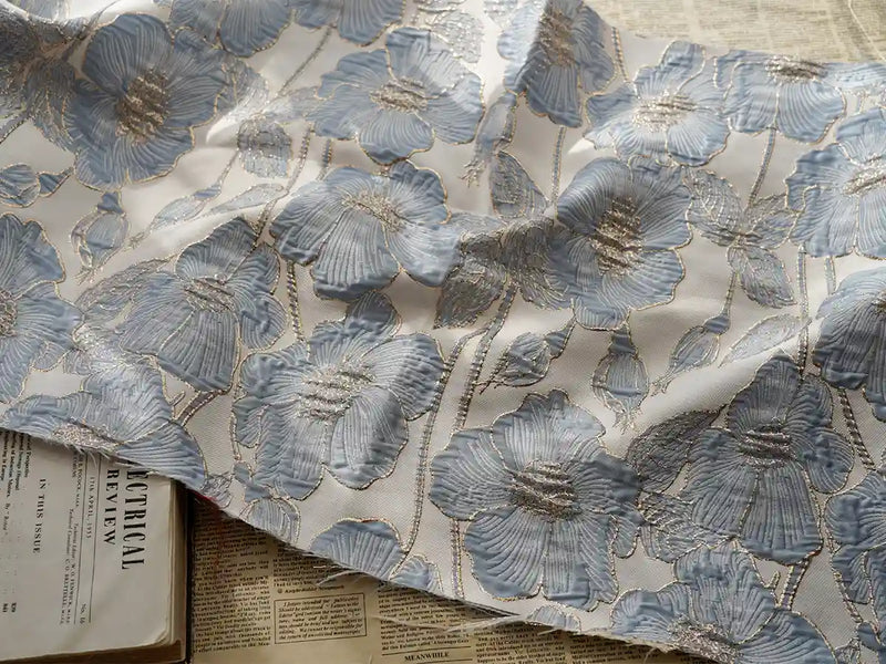 Un morceau de Tissu brocart floral 3D en fil d'or de Longan Craft France est posé sur des journaux ouverts, son élégant motif jacquard bleu, argenté et doré apportant une touche de luxe haute couture au-dessus des titres visibles en dessous.