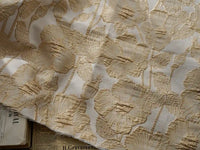 Un gros plan du Tissu brocart floral 3D en fil d'or de Longan Craft France montre une broderie florale dorée complexe sur un jacquard blanc, avec des touches de journaux imprimés en dessous pour un accent sophistiqué et élégant.