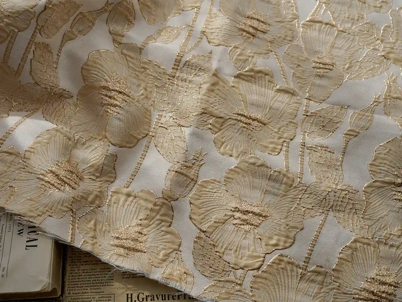 Un gros plan du Tissu brocart floral 3D en fil d'or de Longan Craft France montre une broderie florale dorée complexe sur un jacquard blanc, avec des touches de journaux imprimés en dessous pour un accent sophistiqué et élégant.