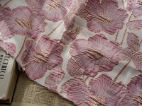 Gros plan sur le tissu brocart floral 3D en fil d'or de Longan Craft France, qui présente un élégant tissu jacquard avec une broderie rose texturée sur un fond clair, drapé sur des pages de livres et de journaux.