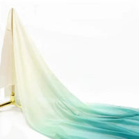 Une Mousseline transparente dégradée au mètre de Longan Craft France en dégradé crème à bleu-vert clair est étalée sur le sol, suspendue à un support sur un fond blanc uni.