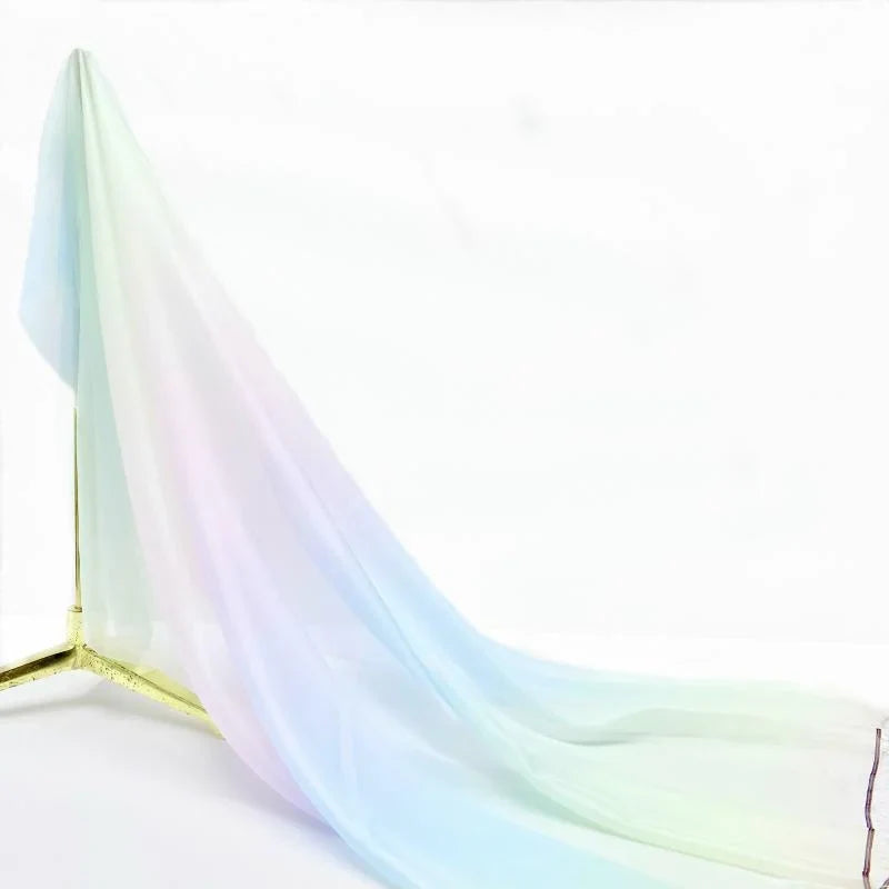 Une Mousseline transparente dégradée au mètre de Longan Craft France est drapée sur un support en métal doré, sur un fond blanc uni, mettant en valeur son délicat dégradé de bleu, de rose et de vert.