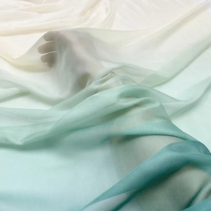 Un pied et une jambe reposent sur une surface douce, partiellement drapée de Mousseline transparente dégradée au mètre de Longan Craft France - un tissu transparent à effet dégradé, blanc et vert pâle, qui confère à la scène un aspect aérien et délicat.