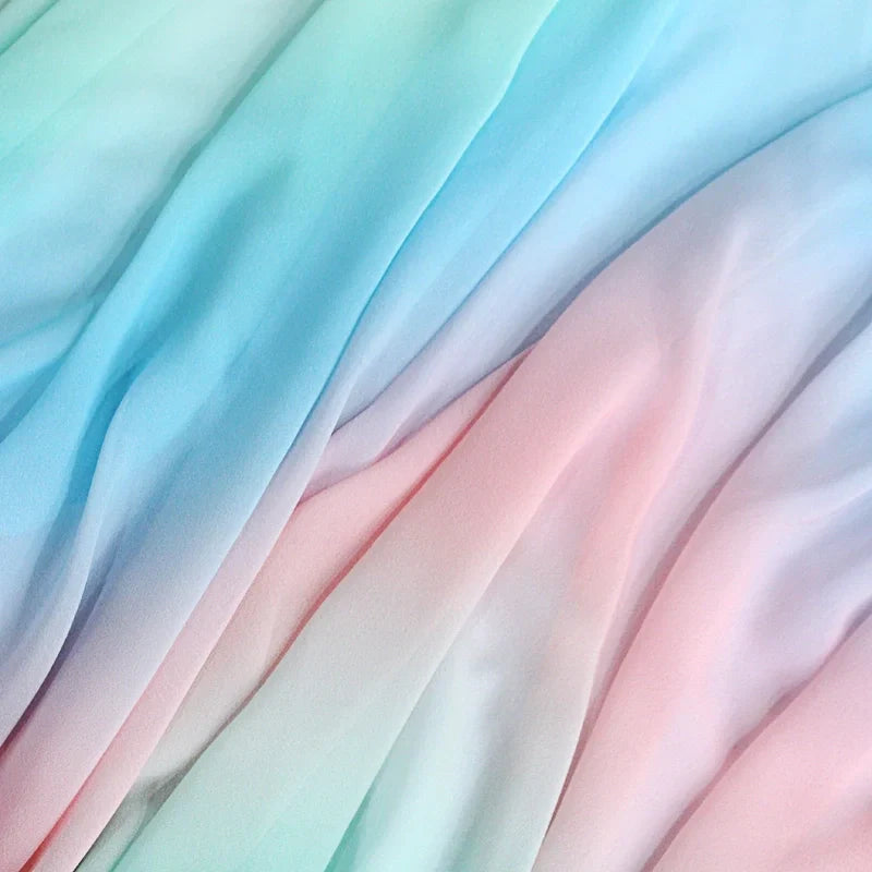 Un gros plan de Tissu en mousseline dégradé arc-en-ciel au mètre de Longan Craft France montre un tissu doux et fluide avec des dégradés pastel - bleu clair, rose et lavande - créant des plis doux et ondulés ; idéal pour les peignoirs, les blouses ou la décoration.