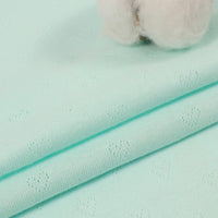 Gros plan sur le tissu jacquard pointelle bleu clair de Longan Craft France, qui présente un délicat motif de cœur, plié en plusieurs couches. Une balle de coton apparaît en haut à droite. Idéal pour les vêtements d'été pour bébés et enfants.