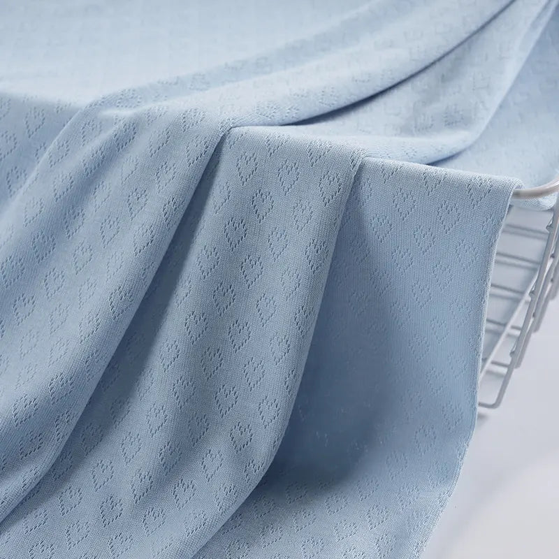 Un gros plan du tissu pointelle cœur au mètre bleu clair de Longan Craft France montre son motif subtil drapé sur un panier en métal. Ce tissu extensible est parfait pour les couches de base et les vêtements d'intérieur DIY, car il offre une douceur et une texture délicate.