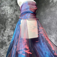 Une robe est drapée de "Tissu organza irisé au mètre | Matière robe en organza cristal" de Longan Craft France en bleu et rose chatoyants, sur un fond gris texturé, le tissu semi-transparent s'écoulant gracieusement jusqu'au sol.