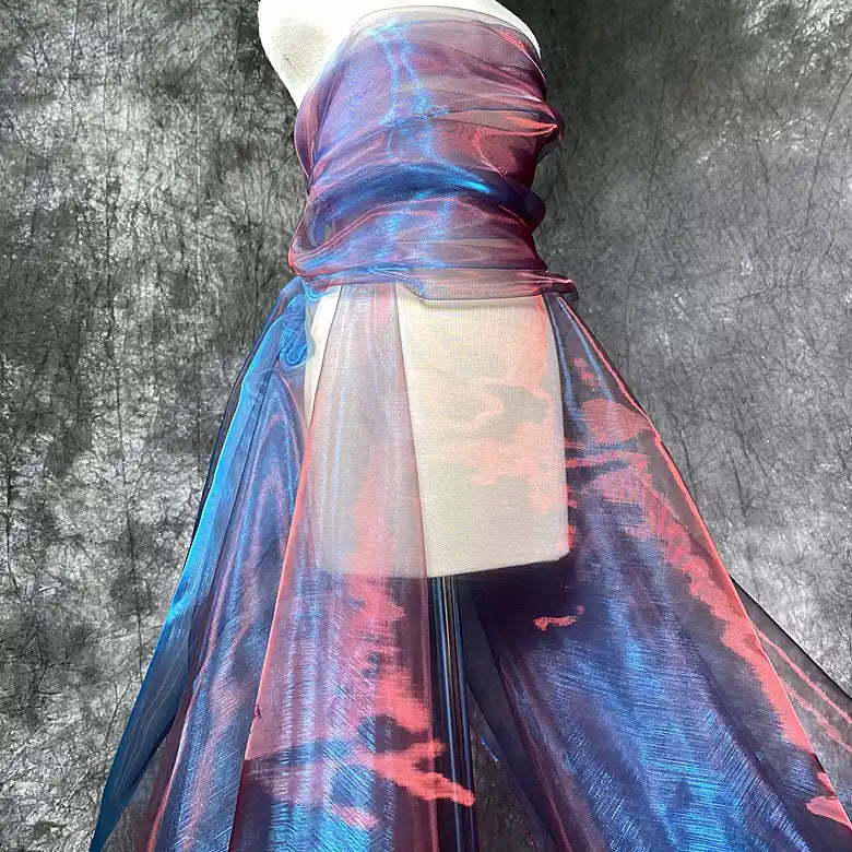 Une robe est drapée de "Tissu organza irisé au mètre | Matière robe en organza cristal" de Longan Craft France en bleu et rose chatoyants, sur un fond gris texturé, le tissu semi-transparent s'écoulant gracieusement jusqu'au sol.