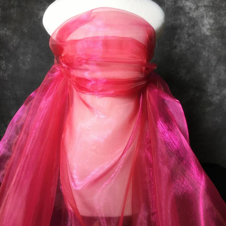 La tête d'un mannequin est drapée de Tissu organza irisé au mètre de Longan Craft France, un tissu organza rose cristal brillant, couvrant la tête et le cou sur un fond texturé sombre.