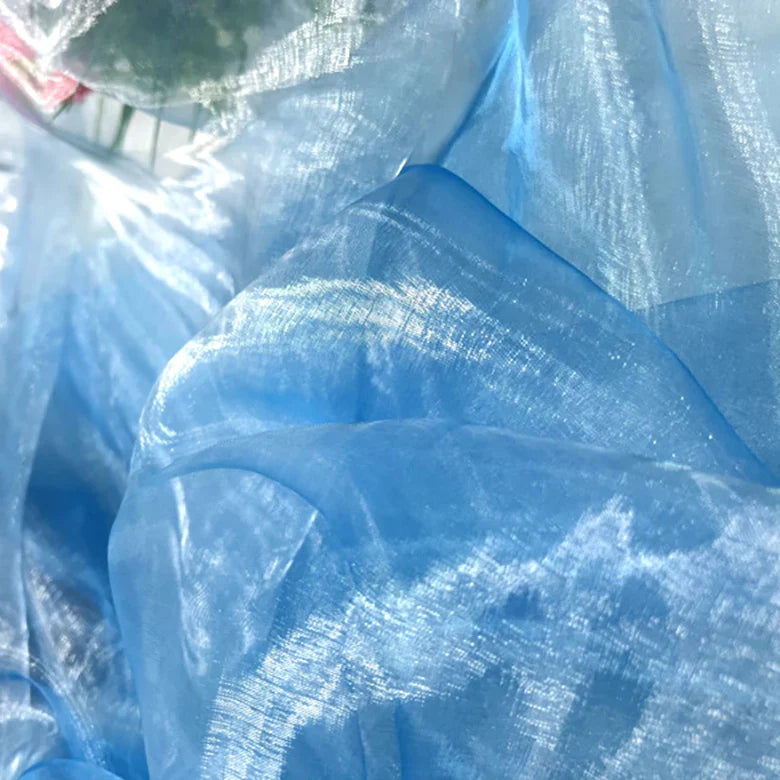 Un gros plan de Tissu organza irisé au mètre de Longan Craft France montre de l'organza cristallin bleu clair avec une texture brillante et froissée reflétant la lumière, et de légères touches de vert et de rose à l'arrière-plan.