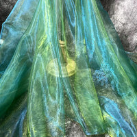 Une main tient un gobelet en laiton, partiellement dissimulé derrière des couches de Tissu organza irisé au mètre de Longan Craft France, drapé sur une surface grise texturée.