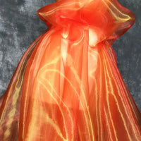 Un "Tissu organza irisé au mètre" orange transparent de Longan Craft France, présentant un subtil motif jaune, est drapé et noué dans un nœud sur un fond texturé gris.