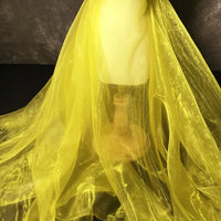 Un Tissu organza irisé au mètre de Longan Craft France, de couleur jaune, se drape sur une robe avec un support en bois, sur un fond sombre. Ce tissu organza cristal est léger, transparent et brillant avec des plis doux.