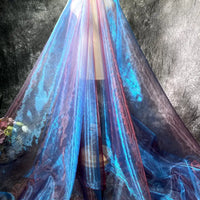 Un mannequin est drapé jusqu'au sol dans le Tissu organza irisé au mètre de Longan Craft France, un organza cristal bleu et rose transparent. À gauche, un panier de fleurs roses et jaunes se détache sur un fond sombre texturé.