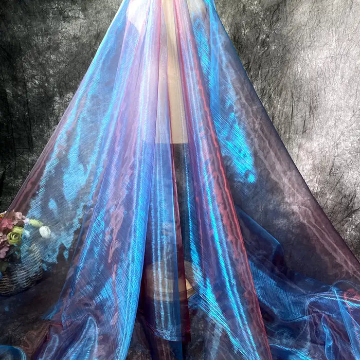 Un mannequin est drapé jusqu'au sol dans le Tissu organza irisé au mètre de Longan Craft France, un organza cristal bleu et rose transparent. À gauche, un panier de fleurs roses et jaunes se détache sur un fond sombre texturé.