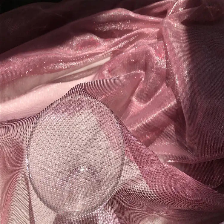Un verre transparent est posé sur le Tissu en tulle bicolore irisé au mètre de Longan Craft France, un tissu chatoyant, semblable à une maille, dont les plis et les reflets irisés sont mis en valeur par la lumière - idéal pour créer des robes éblouissantes.