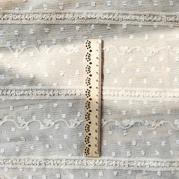 Une règle en bois est posée verticalement sur le tissu en mousseline jacquard à rayures et pois au mètre de Longan Craft France, un tissu beige avec des motifs de dentelle et de pointillés, idéal pour les vêtements aérés et les décors élégants. La règle indique des mesures en centimètres.