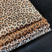 Quatre pièces de Suédine imprimée léopard de Longan Craft France, présentant un imprimé léopard marron, beige et noir, sont empilées sur une surface noire. Ce tissu imitation daim est parfait pour les projets de bricolage de chapeaux, de chaussures et de vêtements.