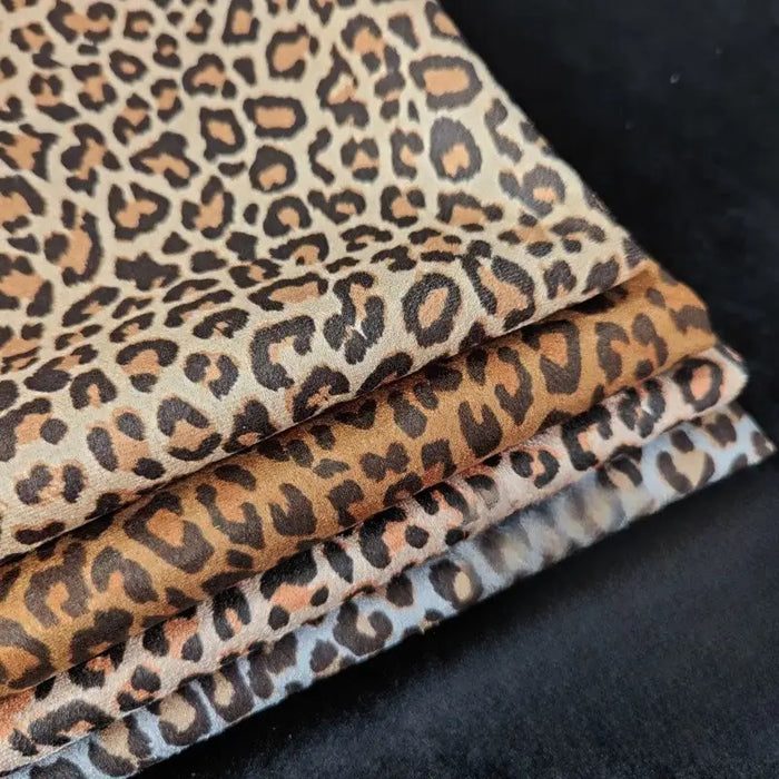 Quatre pièces de Suédine imprimée léopard de Longan Craft France, présentant un imprimé léopard marron, beige et noir, sont empilées sur une surface noire. Ce tissu imitation daim est parfait pour les projets de bricolage de chapeaux, de chaussures et de vêtements.