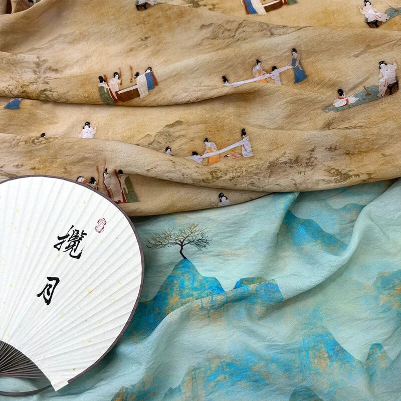 Un éventail décoratif avec des caractères chinois repose sur le tissu en lin imprimé vintage style chinoiserie au mètre de Longan Craft France ; le dessus est orné de petits personnages peints dans des vêtements traditionnels, tandis que le dessous bleu clair montre un paysage et un arbre peints.