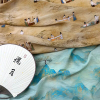 Un éventail décoratif avec des caractères chinois repose sur le tissu en lin imprimé vintage style chinoiserie au mètre de Longan Craft France ; le dessus est orné de petits personnages peints dans des vêtements traditionnels, tandis que le dessous bleu clair montre un paysage et un arbre peints.