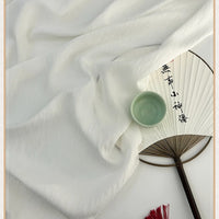 Un tissu lyocell en lin de style chinoiserie vintage de Longan Craft France est drapé à côté d'un éventail en papier à caractères chinois et surmonté d'un petit bol en céramique verte, tandis qu'un pompon rouge apparaît au bas de l'étagère.