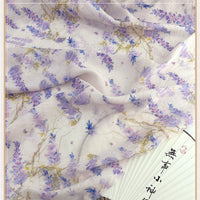 Un tissu chinois vintage en lin-lyocell de Longan Craft France, avec des fleurs violettes et bleues et des vignes vertes, est drapé sur un éventail en papier blanc avec des caractères chinois noirs.