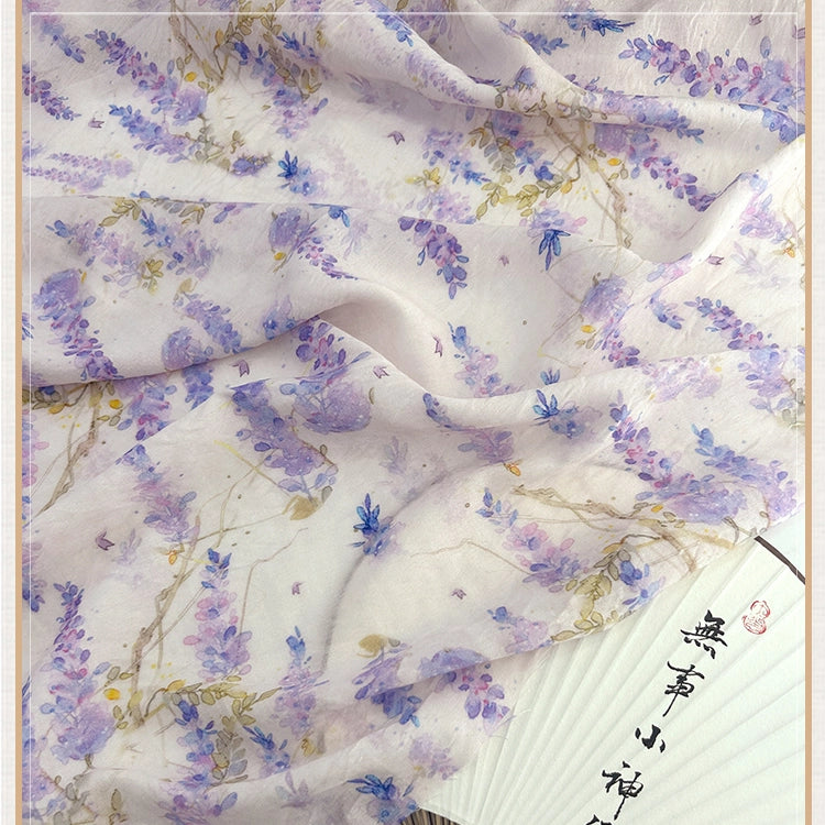 Un tissu chinois vintage en lin-lyocell de Longan Craft France, avec des fleurs violettes et bleues et des vignes vertes, est drapé sur un éventail en papier blanc avec des caractères chinois noirs.