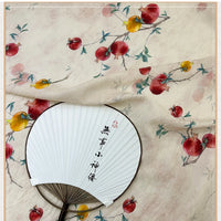 Un éventail rond avec des calligraphies chinoises est posé sur du Tissu en lin imprimé vintage style chinoiserie au mètre par Longan Craft France, avec un motif de branche de grenade avec des fruits rouges et jaunes, des feuilles vertes, sur du tissu lyocell en lin clair.