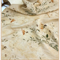 Un morceau légèrement froissé du tissu en lin imprimé vintage style chinoiserie au mètre de Longan Craft France présente des abeilles, des papillons et des branches vertes feuillues, drapés nonchalamment sur la surface.