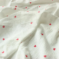Gros plan sur le Tissu double regard petit cœur au mètre de Longan Craft France, un crêpe 100 % coton à la texture froissée, avec des motifs de cœurs rouges régulièrement espacés.
