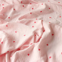 Tissu double gaze petit cœur au mètre de Longan Craft France : crêpe 100% coton, imprimé de petits cœurs rouges sur fond rose pâle.