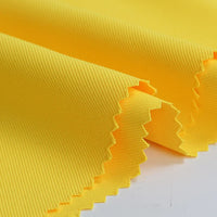 Gros plan de deux pièces superposées de Tissu gabardine à finition mate au mètre de Longan Craft France, montrant la texture sergée nervurée jaune vif et les bords coupés en zigzag, posées à plat sur une surface blanche.