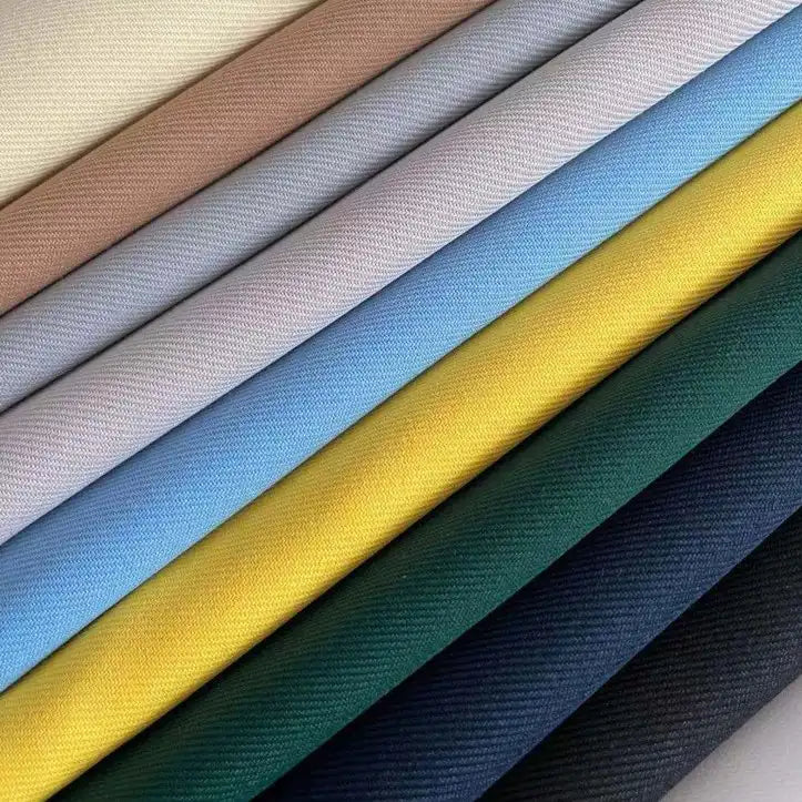 Plusieurs rouleaux de gabardine de qualité supérieure à finition mate de Longan Craft France sont empilés en diagonale dans des tons unis de beige, rose, blanc, bleu clair, jaune, vert, bleu marine et noir. La texture fine du tissage en sergé est clairement visible.