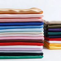 Une pile de Longan Craft France Tissu satin mat uni au mètre dans des couleurs unies - beige, rose, bleu, rouge, vert et blanc - avec d'autres piles de tissus à l'arrière-plan sur la droite.