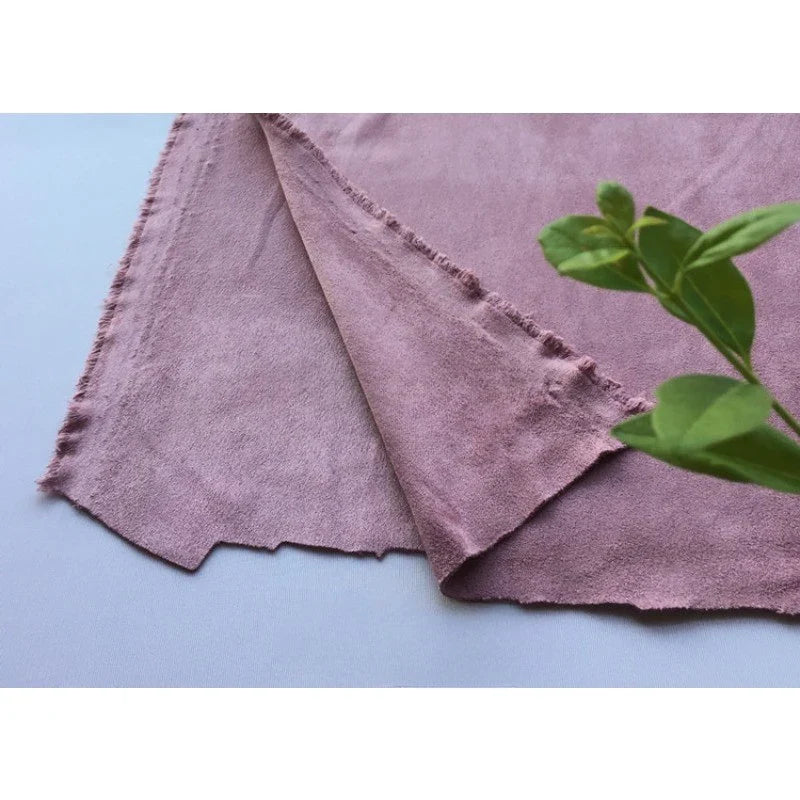 Un morceau de microfibre mauve Matière suédine au mètre de Longan Craft France aux bords rugueux est posé sur une surface plane, une plante à feuilles vertes étant partiellement visible au premier plan. Tissu doux, extensible dans les 4 sens, idéal pour les projets de bricolage.