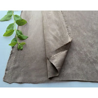 Un morceau de Matière suédine microfibre au mètre de Longan Craft France, de couleur marron clair, aux bords rugueux, est posé à plat à côté d'une petite branche verte feuillue. Ce tissu micro suédine extensible 4 directions est parfait pour les projets de bricolage de vêtements.