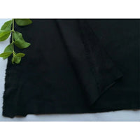 Un morceau de Matière suédine microfibre au mètre de Longan Craft France, de couleur noire, est posé à plat sur une surface blanche, une tige verte feuillue en haut à gauche. Une partie de ce tissu doux et extensible est pliée pour montrer le dessous. Idéal pour les projets de bricolage de vêtements.