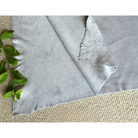 Un morceau de Matière suédine microfibre au mètre de Longan Craft France, un tissu doux et extensible dans les quatre sens pour les vêtements de bricolage, de couleur gris clair, repose sur une surface beige avec des feuilles vertes à proximité ; ses bords sont légèrement inégaux et un coin est plié.