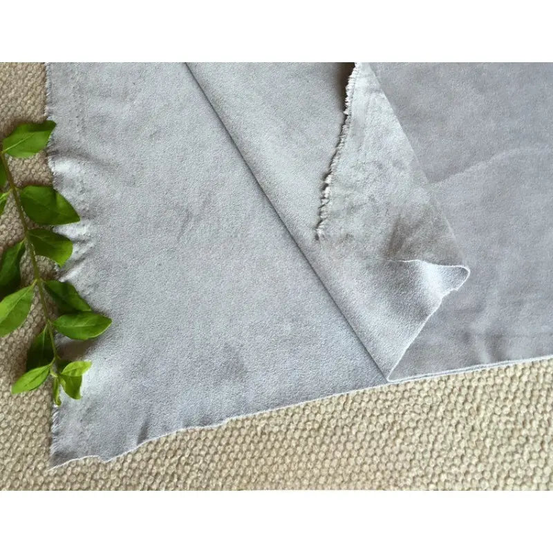 Un morceau de Matière suédine microfibre au mètre de Longan Craft France, un tissu doux et extensible dans les quatre sens pour les vêtements de bricolage, de couleur gris clair, repose sur une surface beige avec des feuilles vertes à proximité ; ses bords sont légèrement inégaux et un coin est plié.