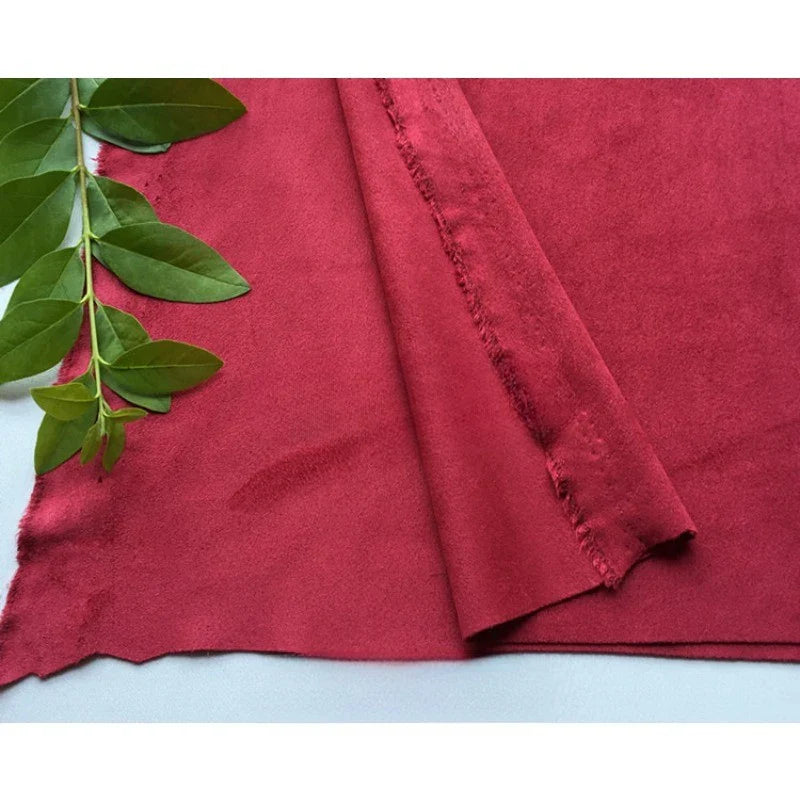 Un morceau de tissu en microfibre suédine de Longan Craft France est plié sur une surface blanche à côté d'une branche verte feuillue. Le tissu micro-suédine est rouge, doux, extensible dans les quatre sens et ses bords ne sont pas finis.