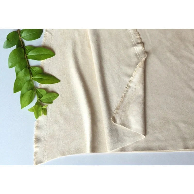 Un morceau de tissu en microfibre suédine de Longan Craft France, écru et blanc cassé, est posé à plat avec des bords effilochés sur une surface blanche. Une branche feuillue verte orne le coin supérieur gauche de ce tissu extensible et doux.