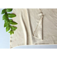Un morceau de tissu en microfibre suédine de Longan Craft France, écru et blanc cassé, est posé à plat avec des bords effilochés sur une surface blanche. Une branche feuillue verte orne le coin supérieur gauche de ce tissu extensible et doux.
