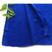 Un gros plan de la matière suédine microfibre au mètre de Longan Craft France, un tissu de bricolage ultra-doux et extensible dans les quatre sens, montre sa couleur bleue vive et son bord brut, partiellement plié avec une brindille verte reposant dans le coin supérieur gauche.