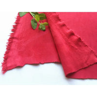 Deux morceaux de "Matière suédine microfibre au mètre" de Longan Craft France, de couleur rouge vif et aux bords effilochés, sont posés sur une surface blanche, accentués par une petite branche verte feuillue reposant sur le tissu doux et extensible.